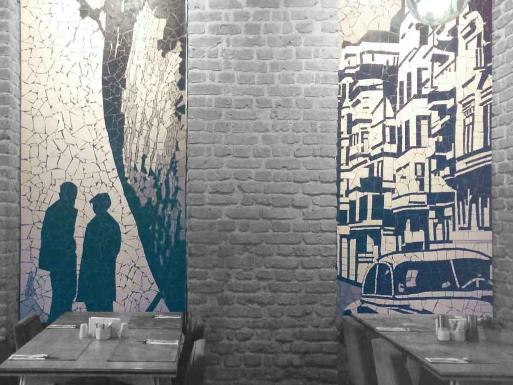 Eine Mosaikwand in einem Restaurant mit 2 Tischen und Besteck im Vordergrund.